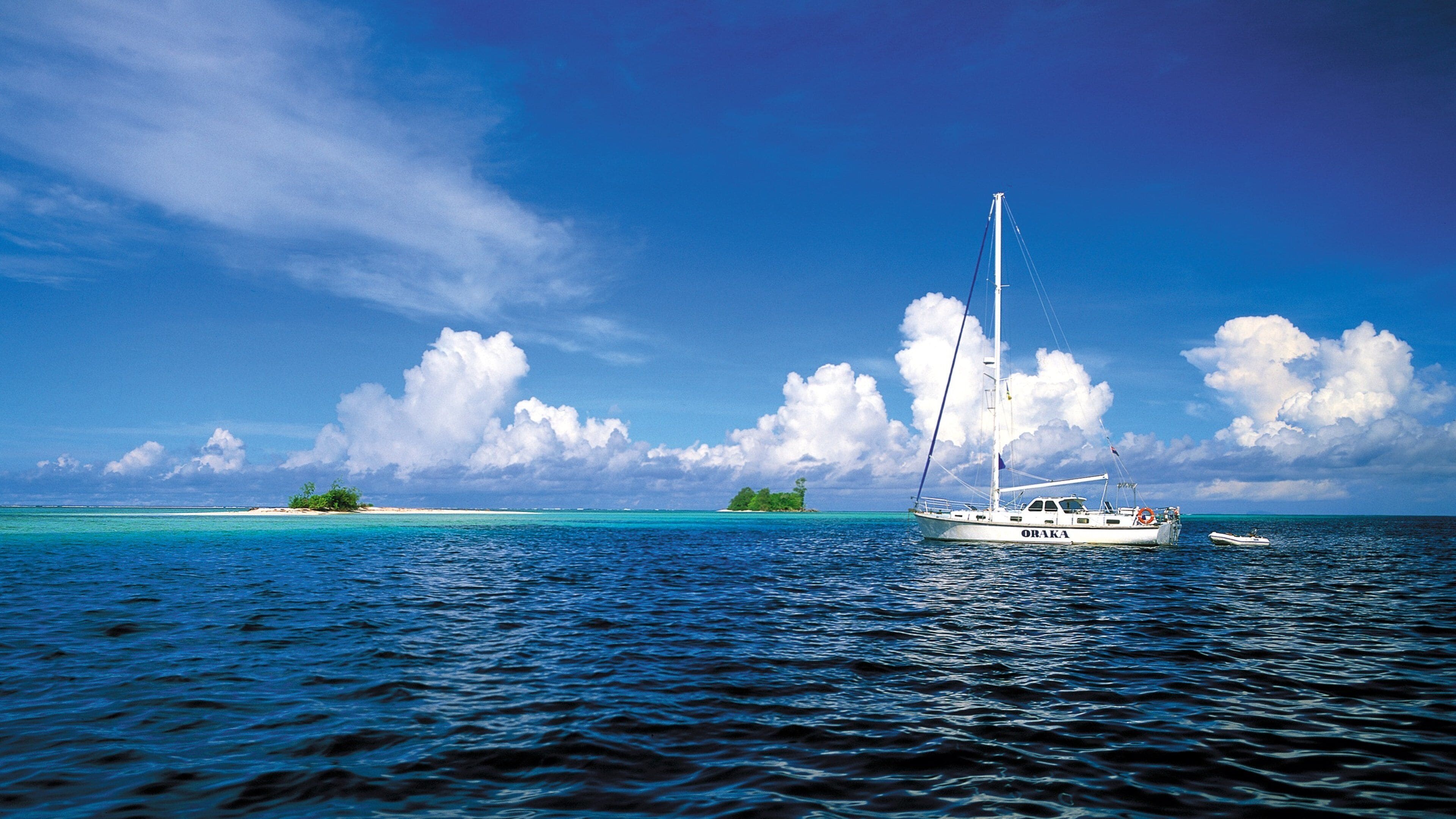 A sailboat named ORAKA on turquoise waters with a small island in the background.