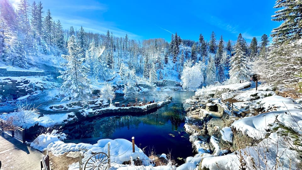 A snow-covered landscape with a clear blue sky, a frozen river, and dense evergreen trees.