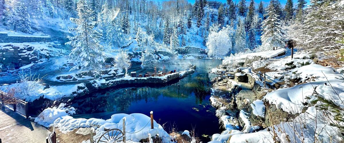A snow-covered landscape with a clear blue sky, a frozen river, and dense evergreen trees.