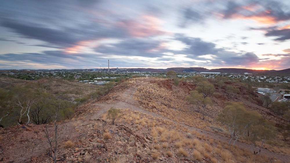 A dry, hilly landscape with sparse vegetation and a distant cityscape under a colorful sunset sky.