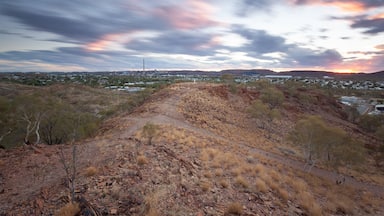 A dry, hilly landscape with sparse vegetation and a distant cityscape under a colorful sunset sky.