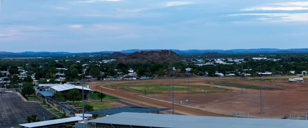 A large open field with solar panels, surrounded by trees and buildings, with mountains in the background.