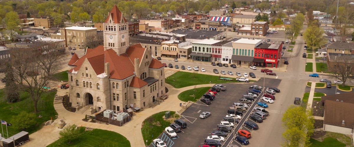 An aerial view of a town with a prominent historic building with a clock tower, a parking lot, and surrounding residential and commercial buildings.