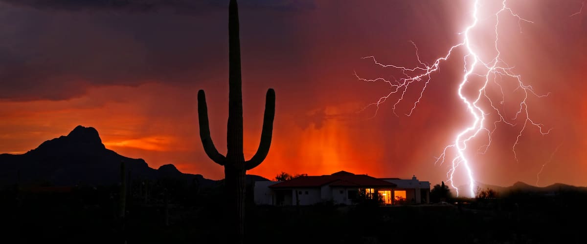 Paisaje desértico con un cactus destacado, una cordillera de montañas y una casa con luces encendidas en su interior.