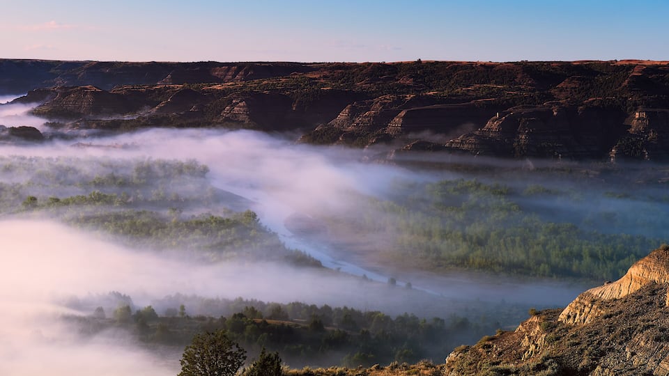 A valley with a river, surrounded by forested hills and a rocky cliff.