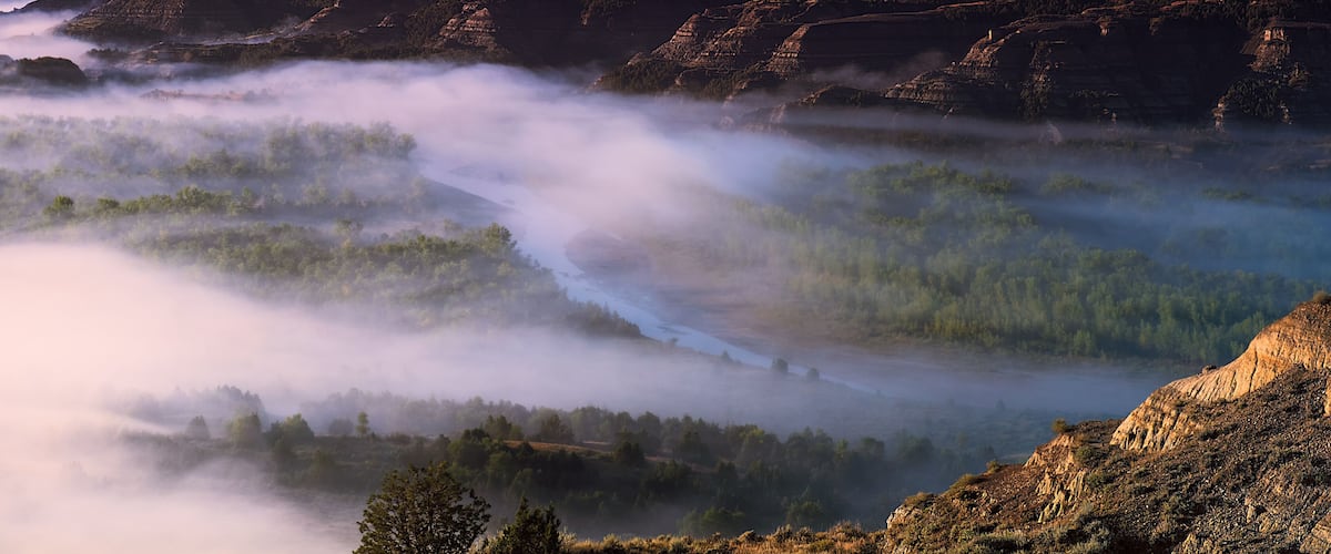 A valley with a river, surrounded by forested hills and a rocky cliff.