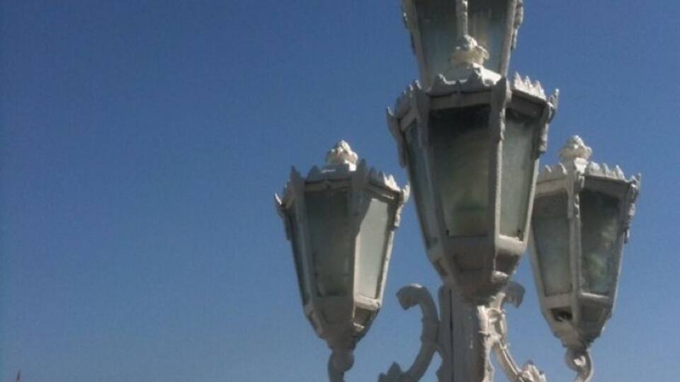 A white ornate street lamp with three lamps, a building with a red roof, and a clear blue sky.