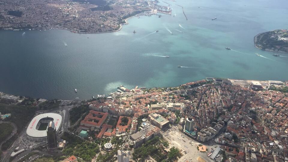 An aerial view of a coastal city with a dense urban center, a large stadium, and a marina with boats.