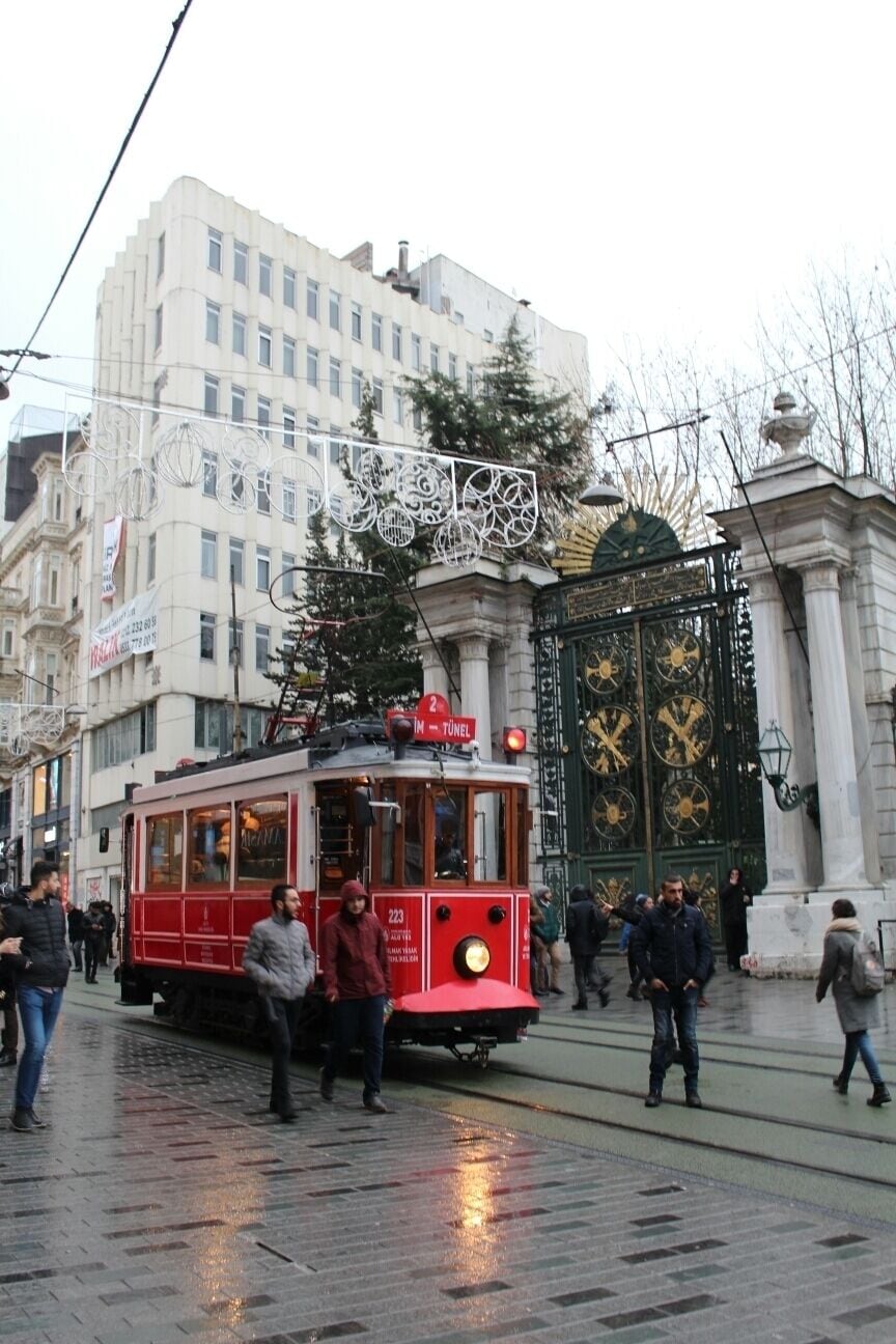 A red tram on a wet street with pedestrians, a large building, and a decorative gate.