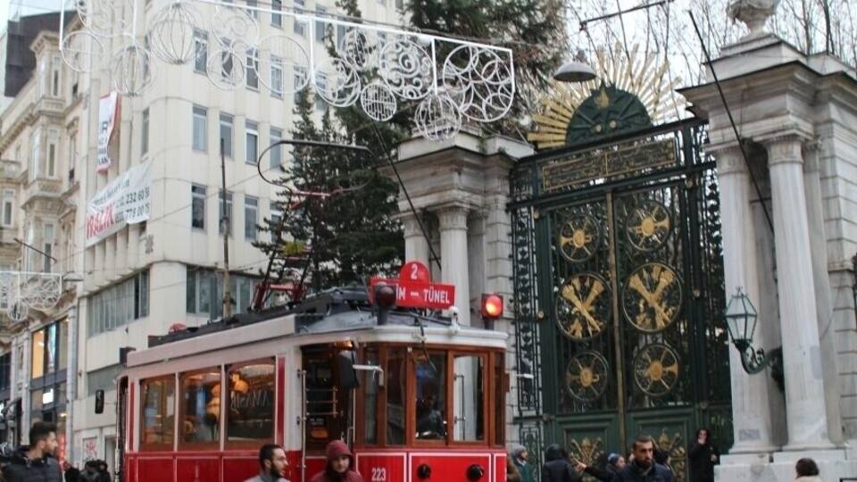 A red tram on a wet street with pedestrians, a large building, and a decorative gate.