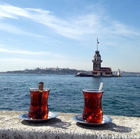 Two glasses of tea on a table with a view of a body of water and a distant building with a tower.