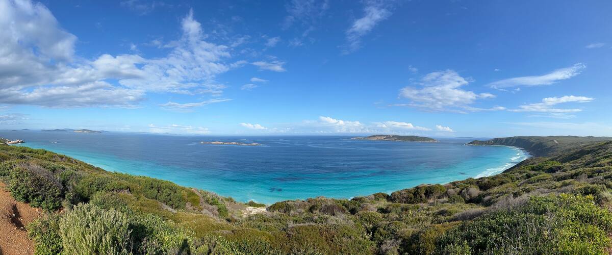 A coastal landscape with a clear blue sea, green hills, and a dirt path.
