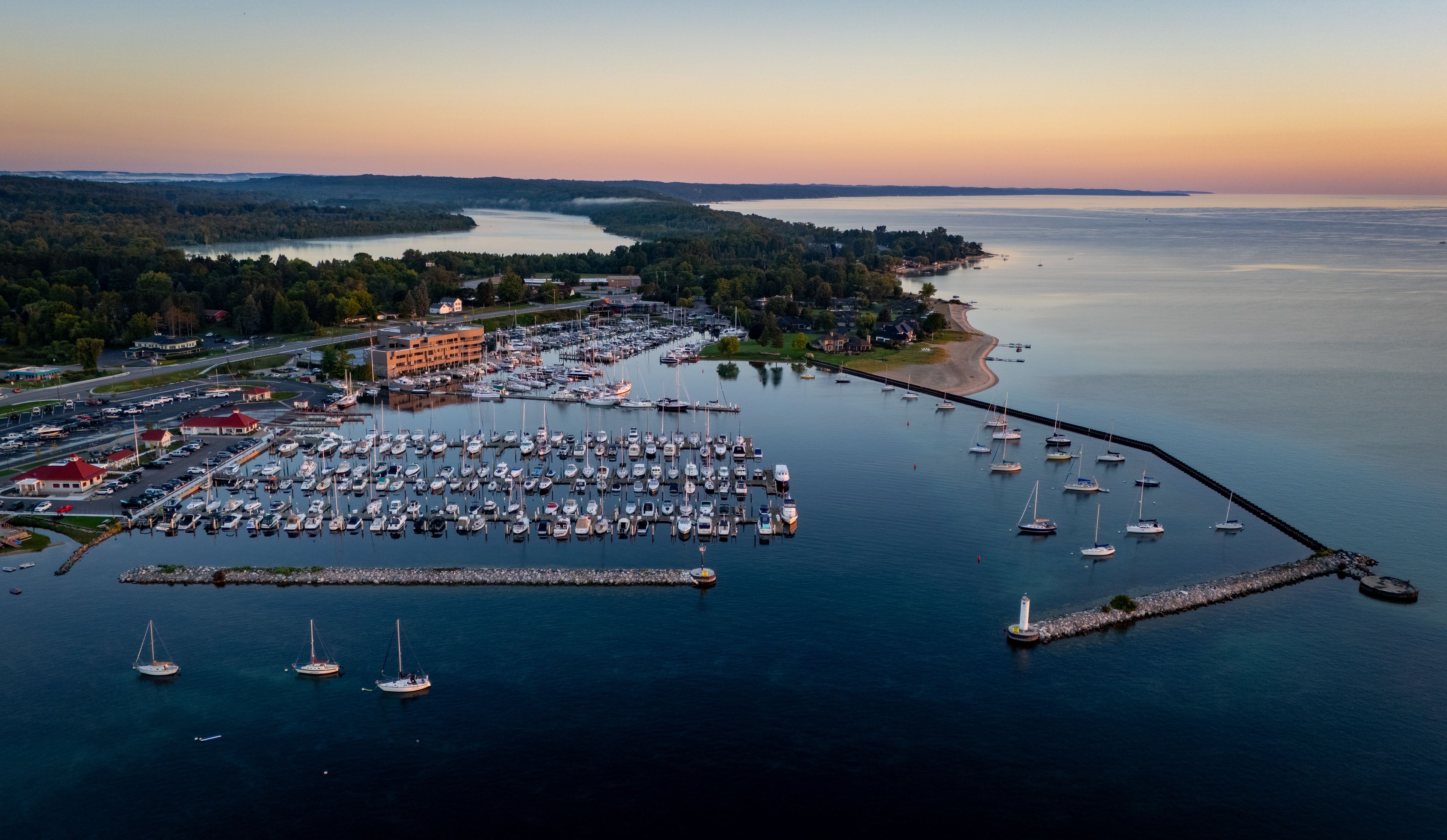 Aerial view of the marina with numerous sailboats docked, a stone breakwater with a lighthouse, and calm water under a golden sunset sky in Traverse City, Michigan, United States.