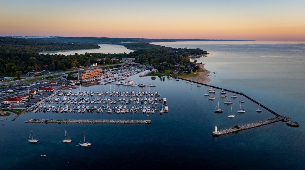 Aerial view of the marina with numerous sailboats docked, a stone breakwater with a lighthouse, and calm water under a golden sunset sky in Traverse City, Michigan, United States.