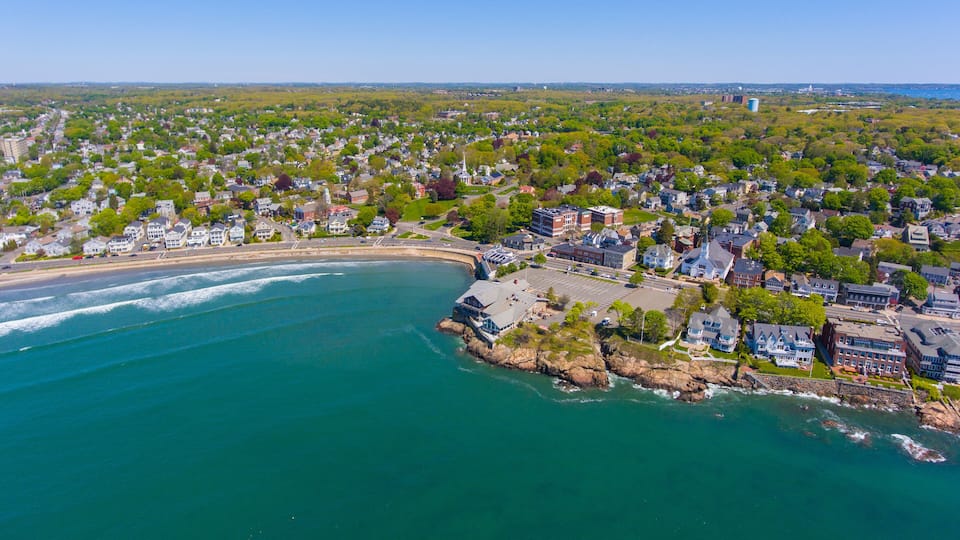An aerial view of a coastal town with a beach, residential areas, and a rocky shoreline.