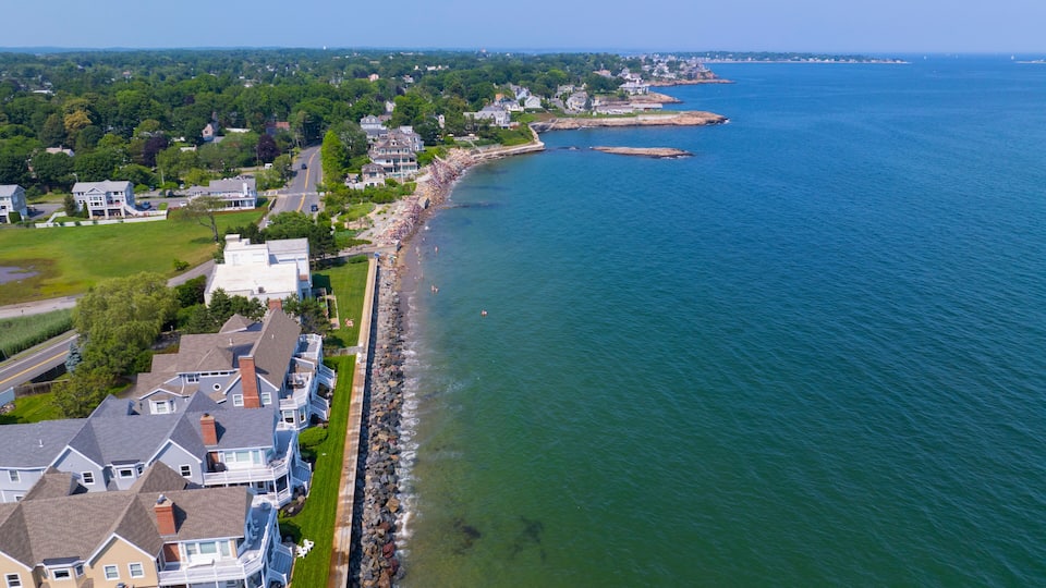 A coastal residential area with houses along the shoreline, a rocky breakwater, and a clear view of the ocean.