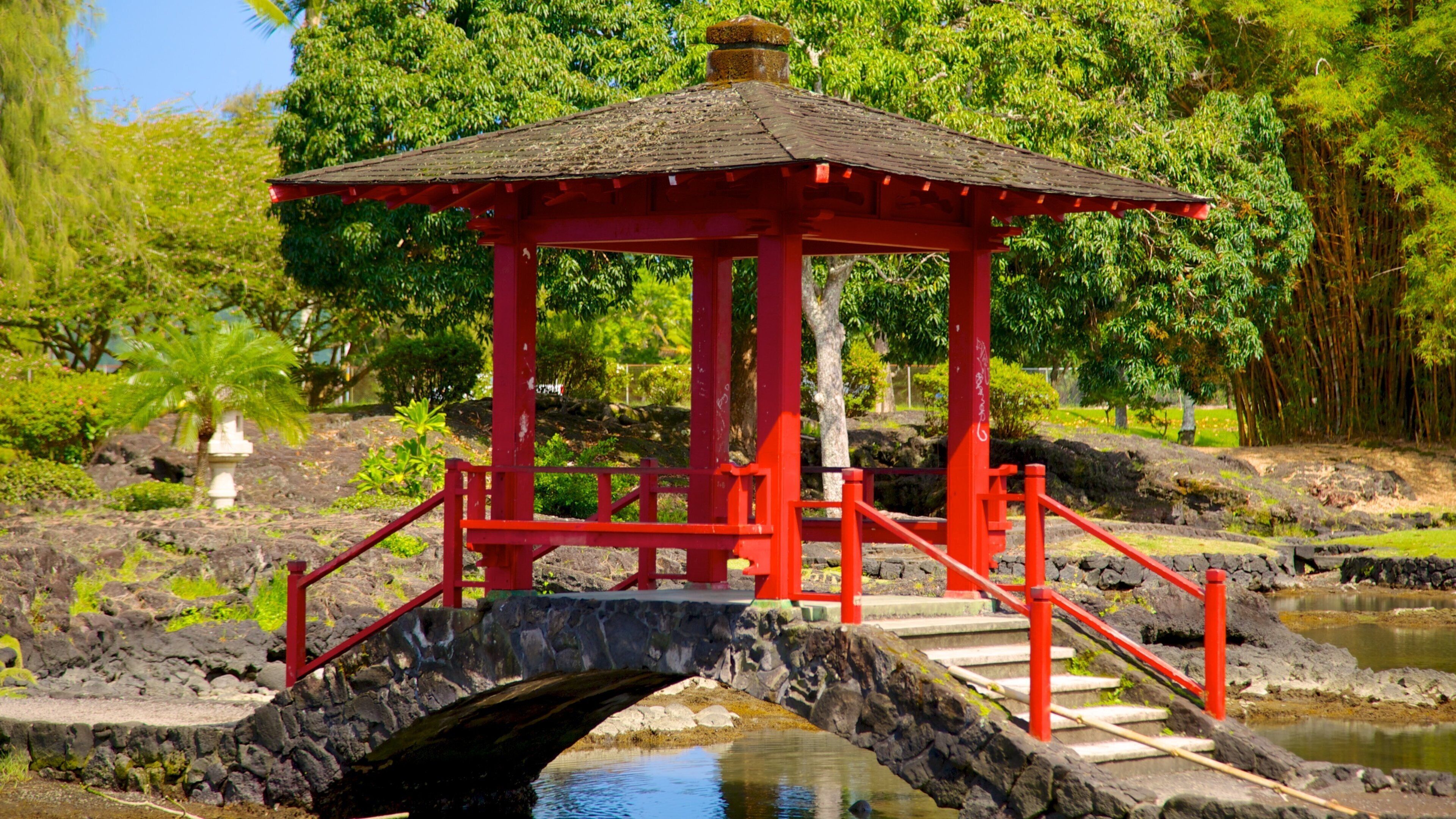 A red gazebo with a stone bridge over a pond, surrounded by lush greenery and trees.