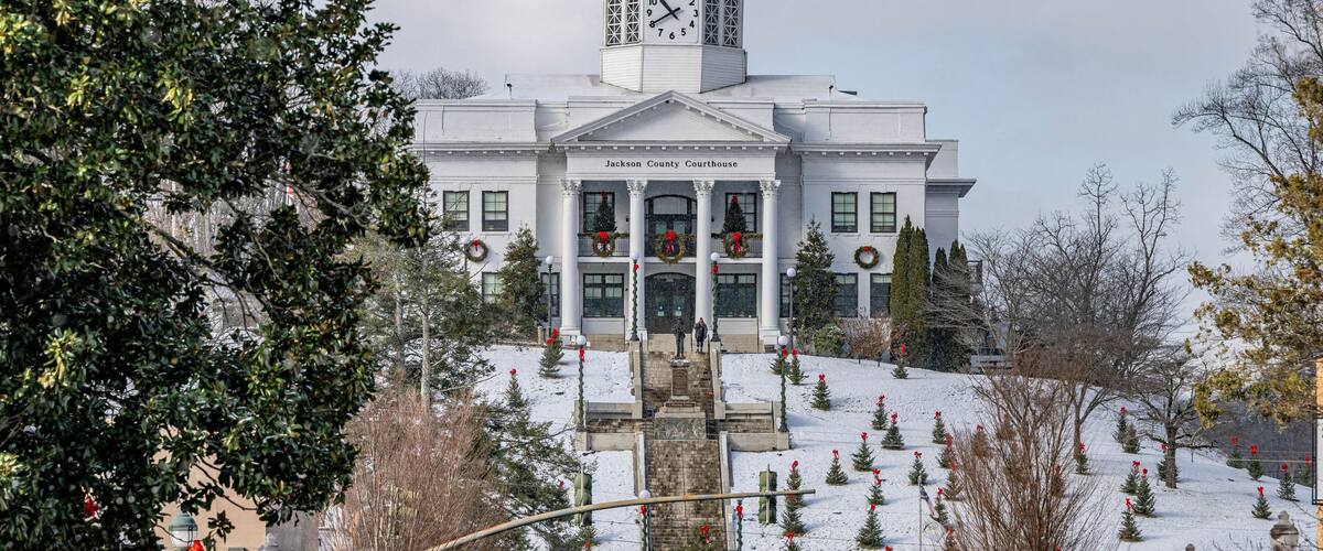 A courthouse with a clock tower, surrounded by snow-covered trees and decorated with wreaths.