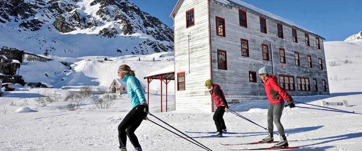 Three skiers on a snowy slope with a wooden building and mountains in the background.