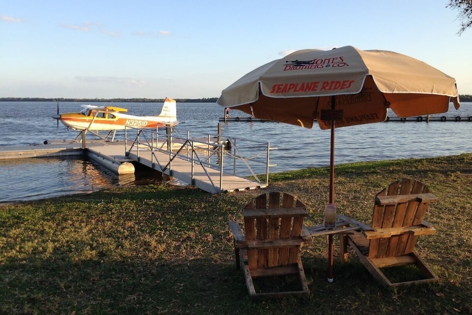 A lakeside setting with a small seaplane docked on the water, a wooden chair and table under a beach umbrella, and a grassy area.