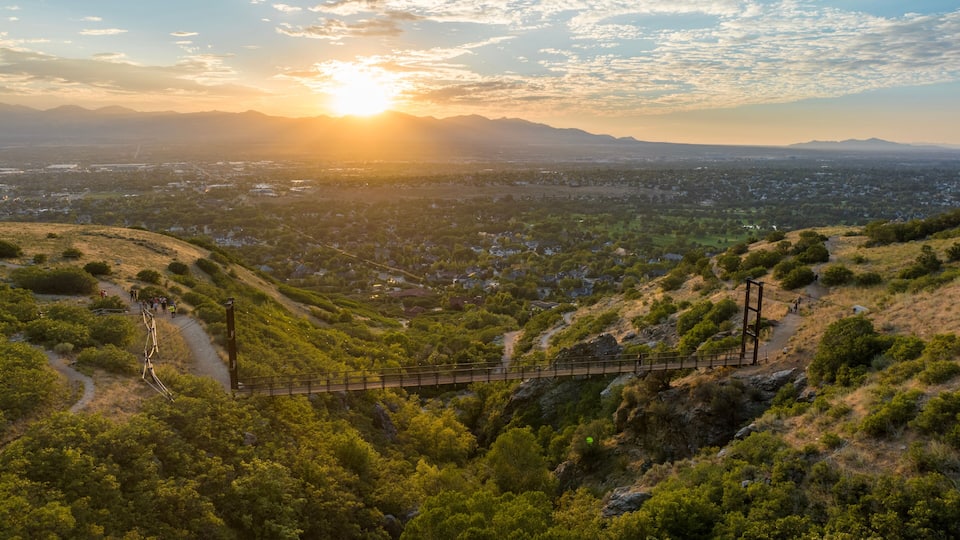 A sunset over a valley with a bridge, surrounded by green hills and a distant cityscape.