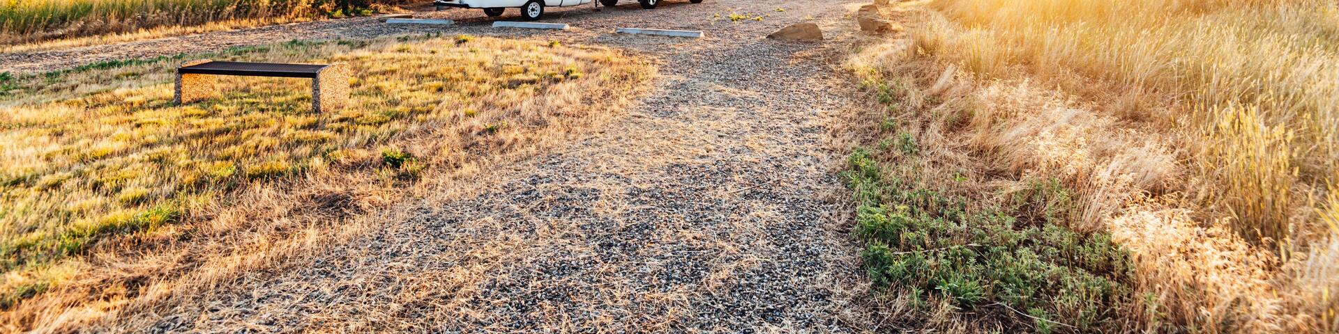 A gravel path leading to a white RV and SUV parked in a grassy field with a sunset in the background.