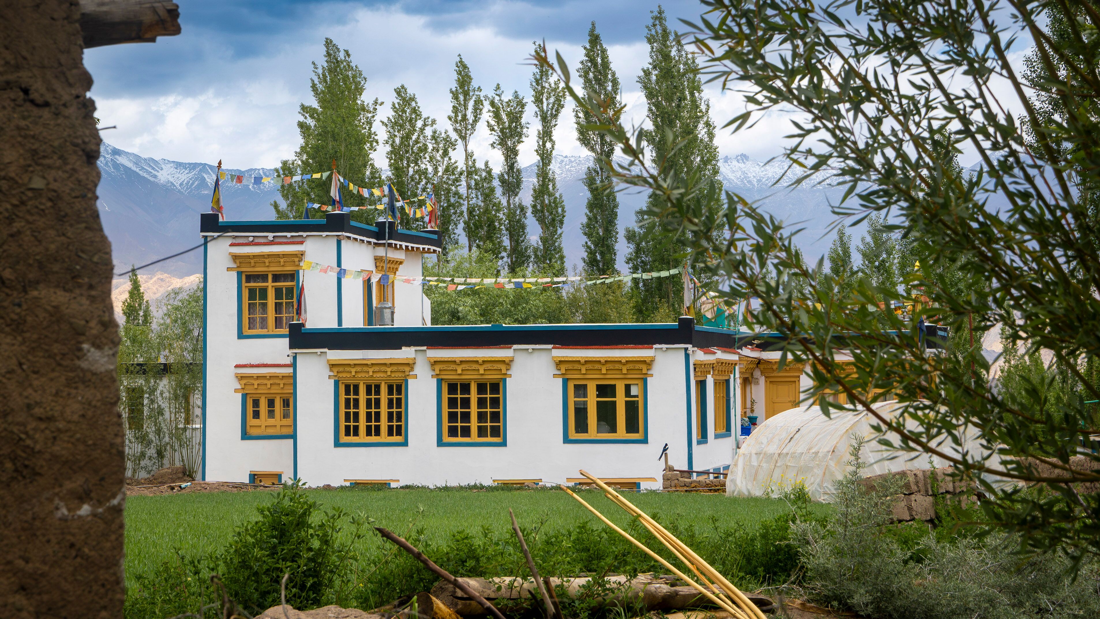 Un edificio tradicional con detalles amarillos y azules, rodeado de vegetación y montañas nevadas al fondo.