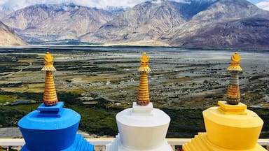 Trois stupas aux couleurs vives, surmontés de dômes dorés, qui dominent une vallée avec des montagnes en arrière-plan.