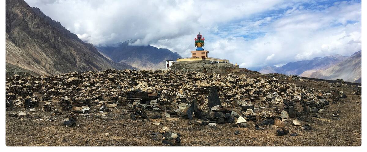 A mountainous landscape with a colorful stupa on a rocky hill, surrounded by rugged terrain and distant mountains under a partly cloudy sky.