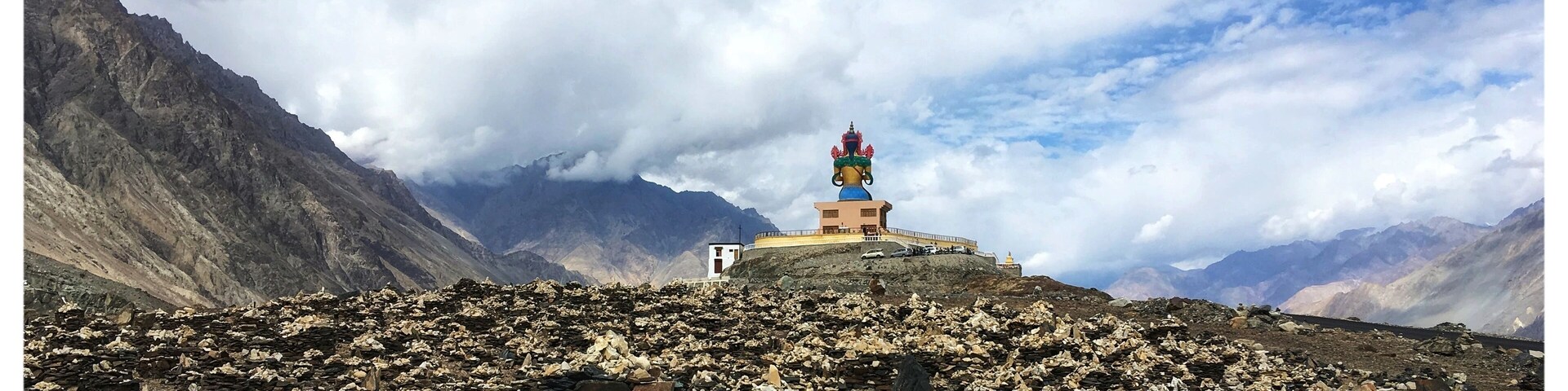 A mountainous landscape with a colorful stupa on a rocky hill, surrounded by rugged terrain and distant mountains under a partly cloudy sky.