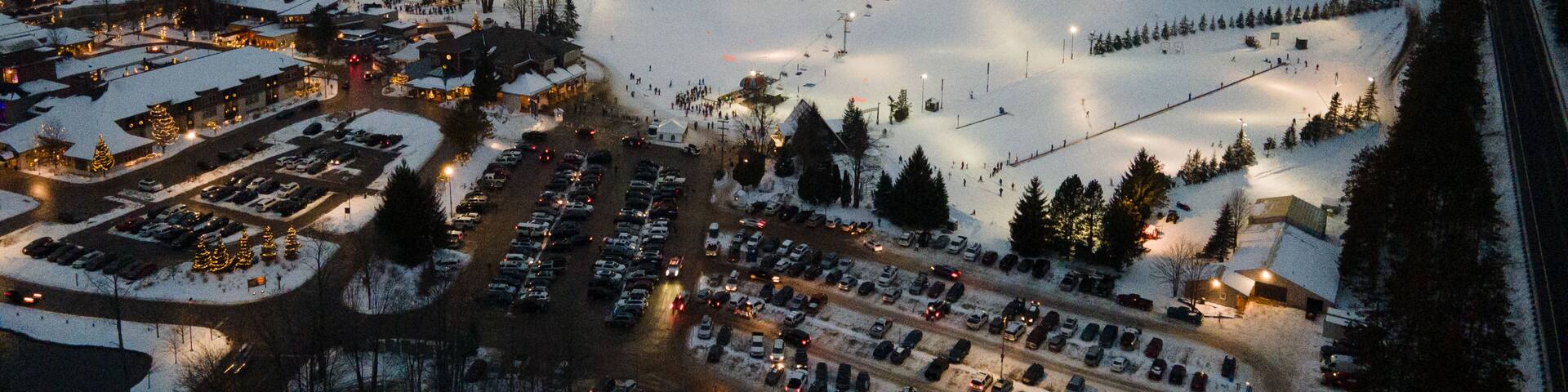 Aerial view of the illuminated ski slopes and base area of Crystal Mountain at night, featuring snow-covered terrain, chairlifts, and a busy parking lot in Thompsonville, Michigan, United States.