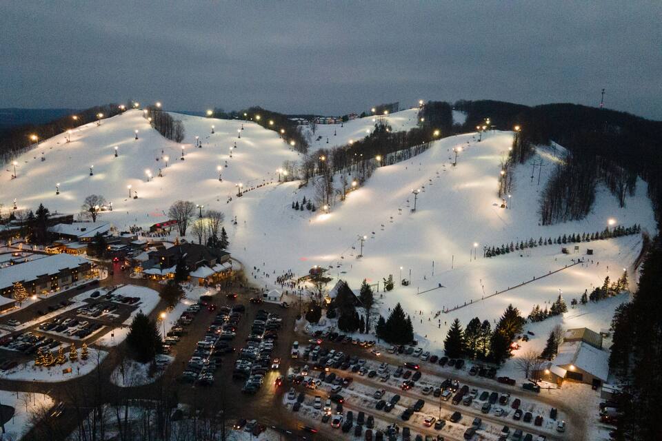 En snötäckt skidort på natten, med upplysta skidliftar och en parkeringsplats.