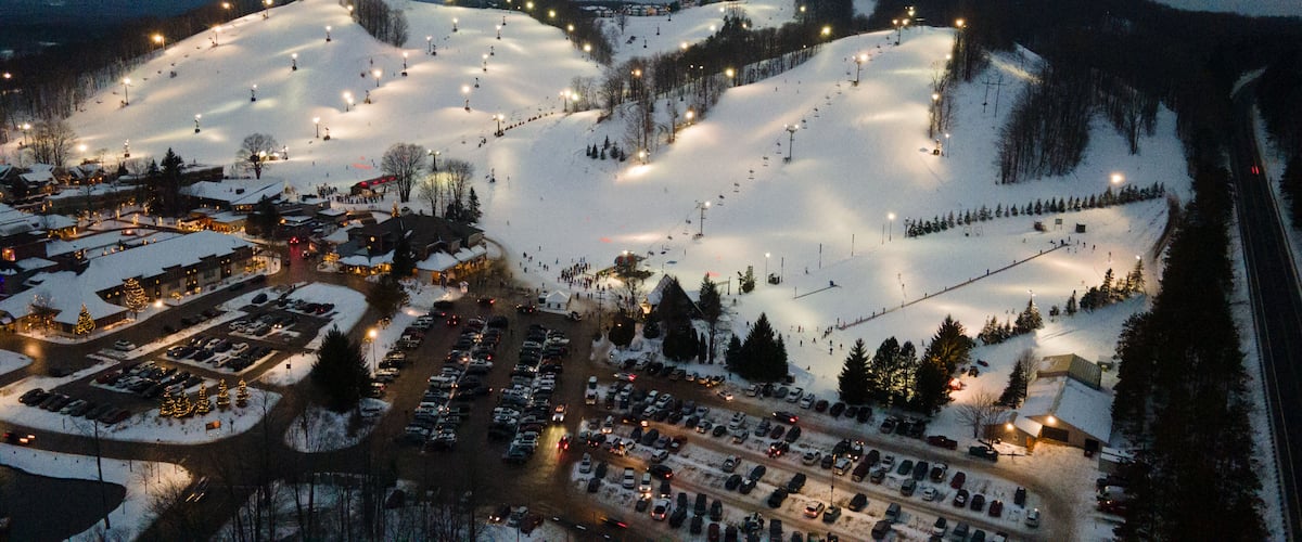 Aerial view of the illuminated ski slopes and base area of Crystal Mountain at night, featuring snow-covered terrain, chairlifts, and a busy parking lot in Thompsonville, Michigan, United States.