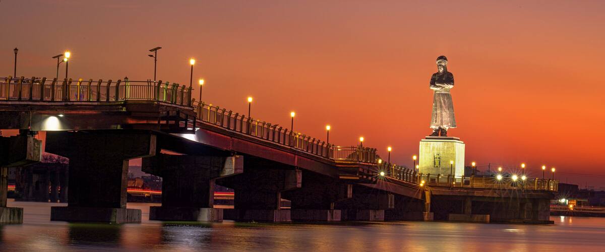 A bridge with a statue at the end, a sunset in the background, and a calm river.