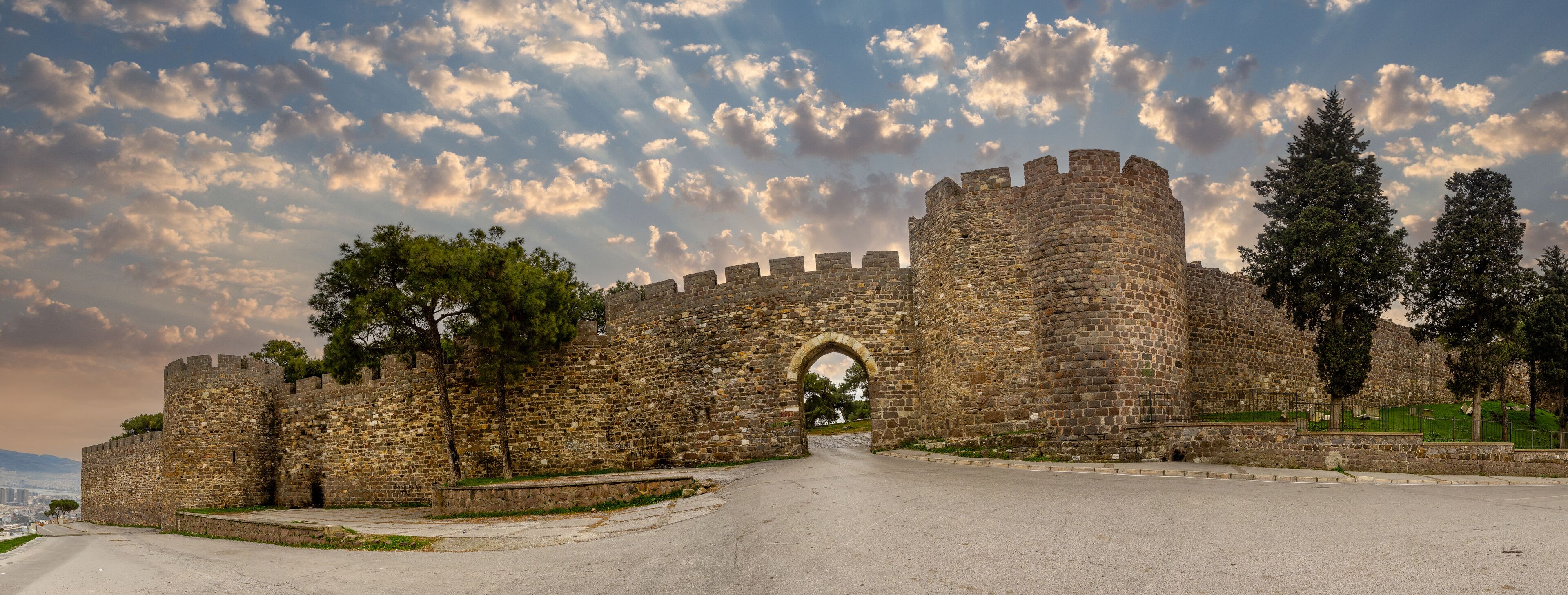 A historic stone castle with a large arched entrance, surrounded by trees and a clear sky with clouds.