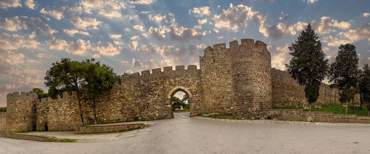 A historic stone castle with a large arched entrance, surrounded by trees and a clear sky with clouds.