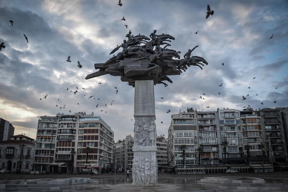 Eine große Säule mit einer Skulptur von Menschen obenauf, umgeben von Vögeln, mit modernen Gebäuden im Hintergrund.