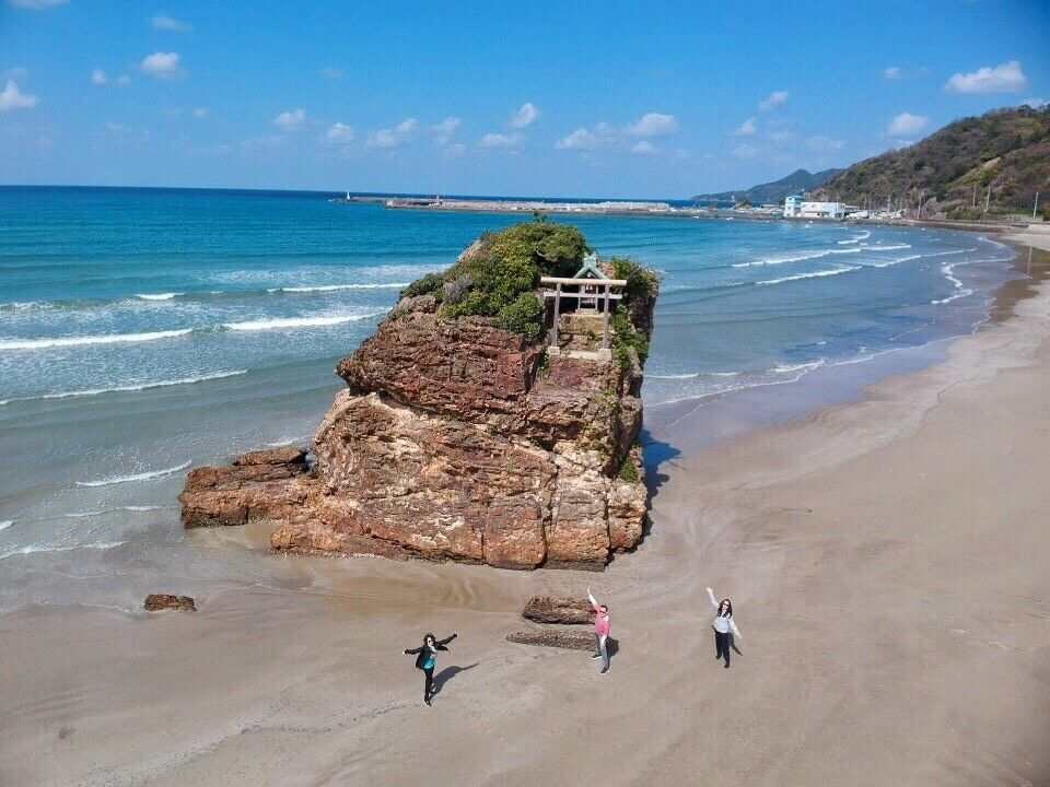 A rocky outcrop with a small structure on a beach, waves crashing, and a clear sky.