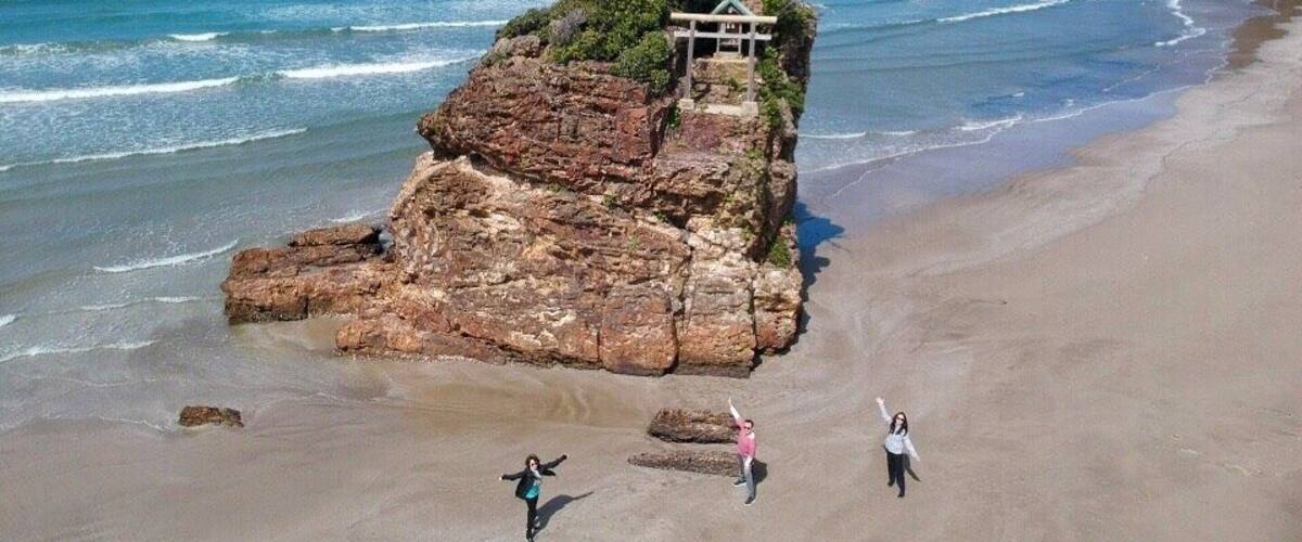 A rocky outcrop with a small structure on a beach, waves crashing, and a clear sky.