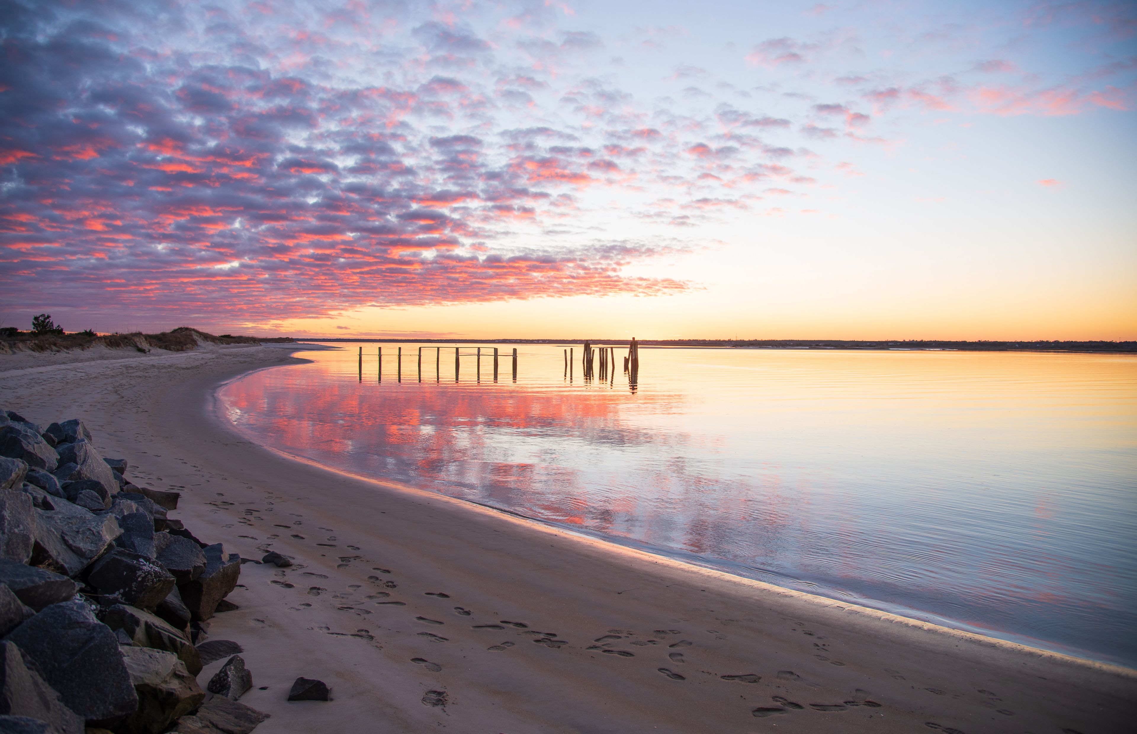 A beach with a wooden pier extending into calm waters, surrounded by rocks and a sunset sky.