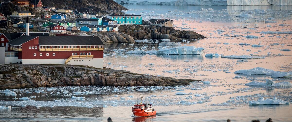 A coastal town with colorful buildings, a red boat, and icebergs in the background.
