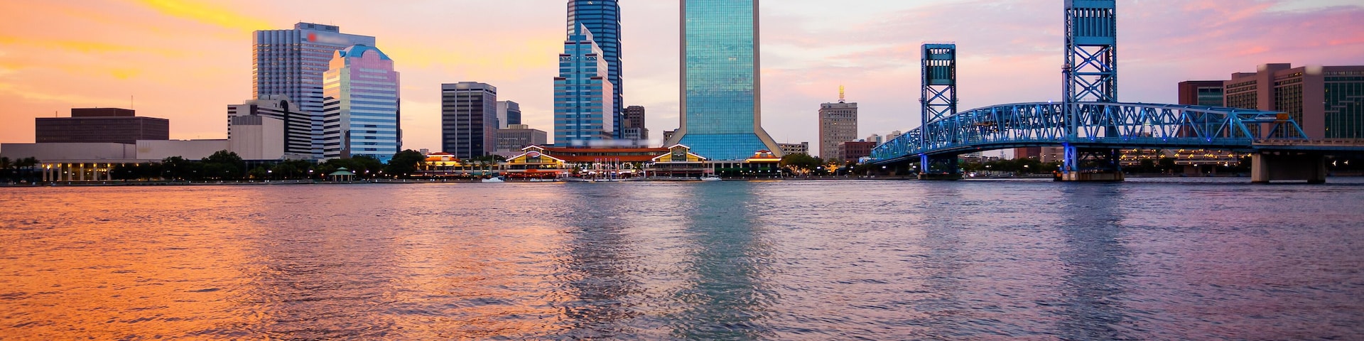 Un horizonte urbano con edificios modernos y un puente azul sobre un río al atardecer.