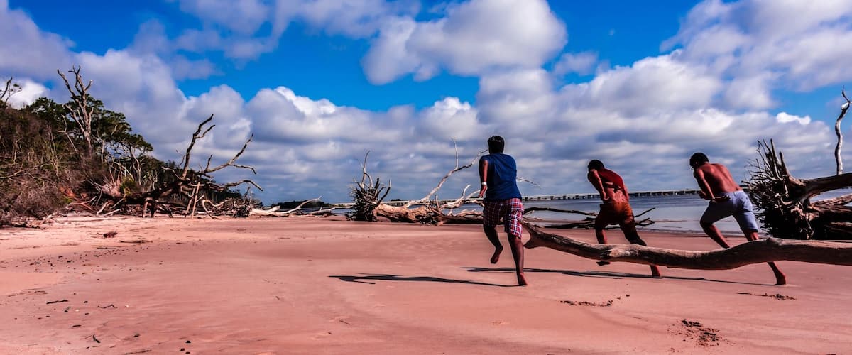A sandy beach with three people playing tug-of-war on a large log.