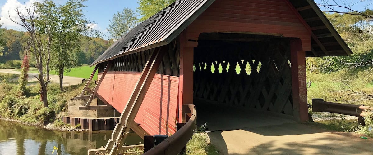 A red covered bridge over a calm river, surrounded by greenery and trees.