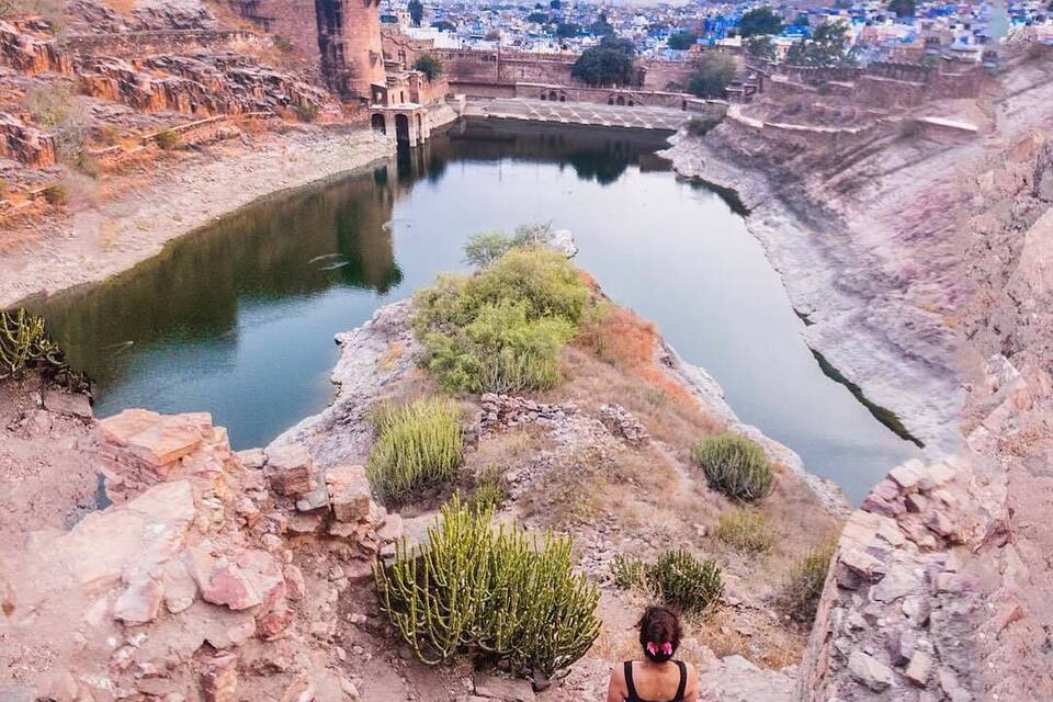 Een persoon loopt over een rotsachtig pad langs een kalme rivier, met een groot fort en een verafgelegen stadsgezicht op de achtergrond.