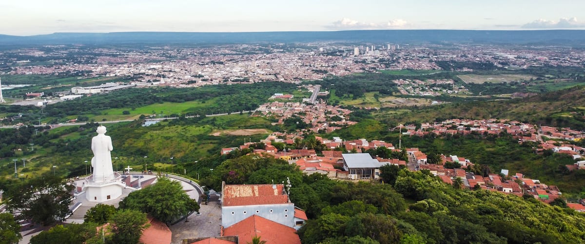 A panoramic view of a city with a prominent white statue, lush green hills, and a mix of residential and commercial buildings.