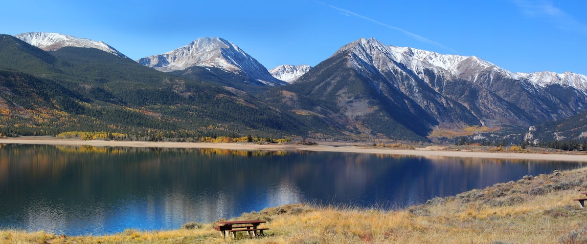 A lake surrounded by mountains with autumn foliage, and two picnic tables on the shore.