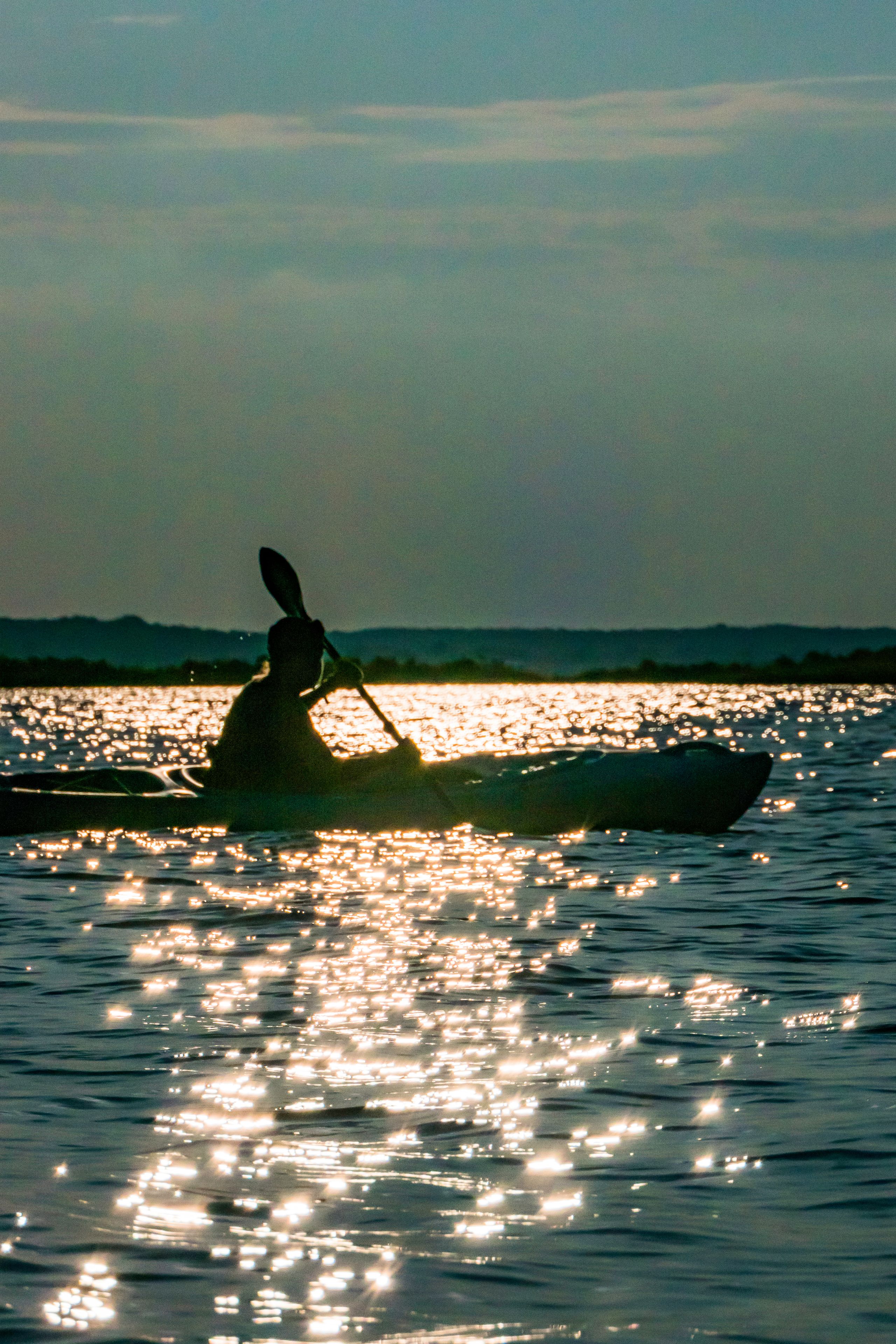 A silhouette of a person kayaking on calm water with the sun reflecting off the surface.