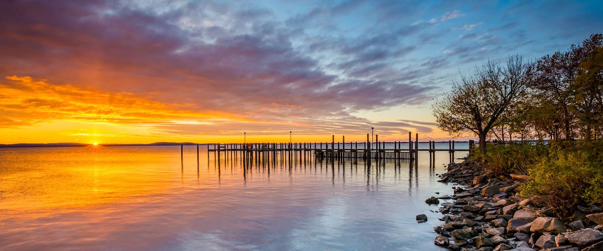 A sunset over a calm lake with a wooden pier extending into the water, surrounded by rocks and trees.