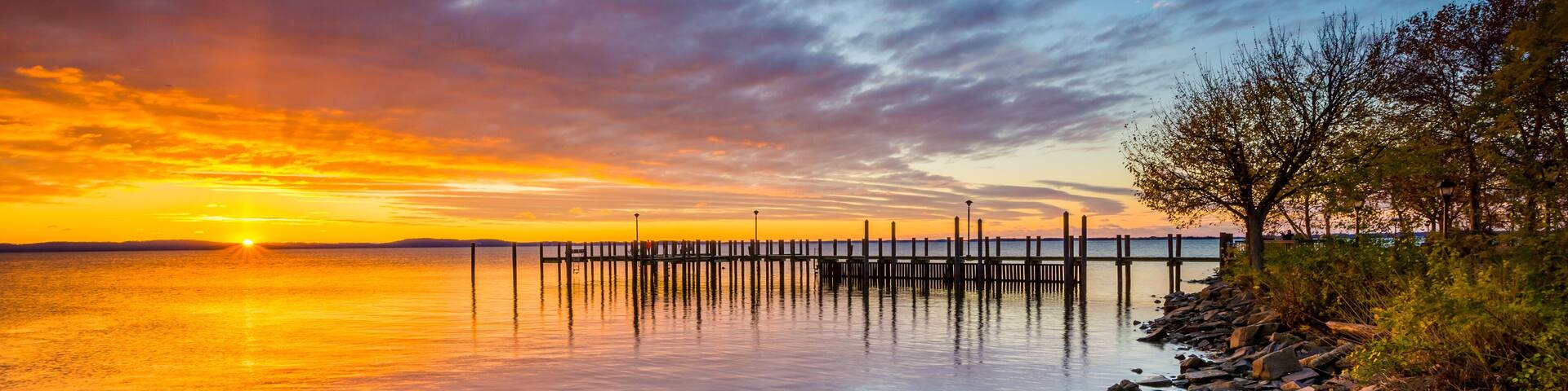 A sunset over a calm lake with a wooden pier extending into the water, surrounded by rocks and trees.